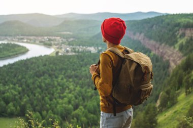 Sırt çantası ve kırmızı şapkalı bir turist uçurumun kenarında duruyor ve nehrin yanındaki yeşil bir vadiye bakıyor. Özgürlük ve doğa ile birlik kavramı.
