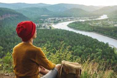 Sırt çantası ve kırmızı şapkalı bir turist uçurumun kenarında oturuyor ve nehrin yanındaki yeşil orman vadisine bakıyor. Özgürlük ve doğa ile birlik kavramı.
