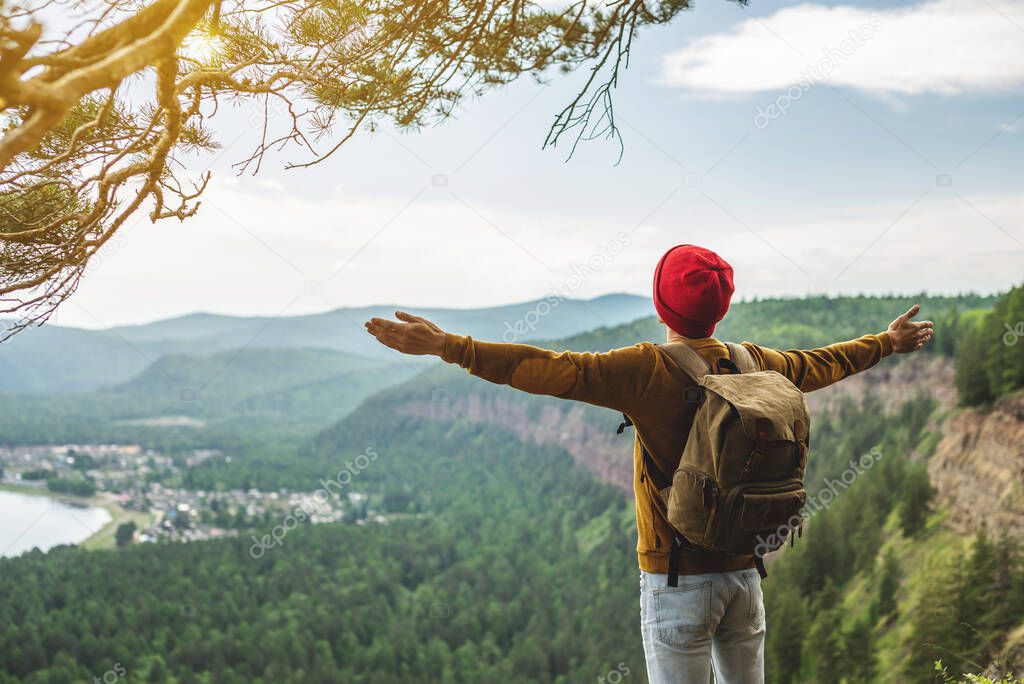A tourist traveler with a backpack and a red hat is standing on the edge of a cliff in front of a green valley with his arms spread wide. The concept of freedom and the joy of success