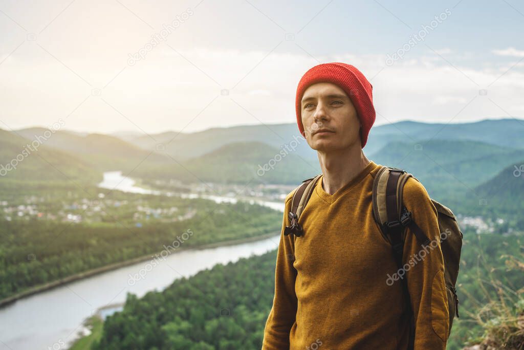 A tourist traveler with a backpack and a red hat is standing on the edge of a cliff and is looking on a green valley with the river. The concept of freedom and unity with nature