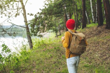 Sırt çantası ve kırmızı şapkalı bir turist ağaçların arasında ormanda yürüyor. Aktif yürüyüş ve doğa ile birlik kavramı.