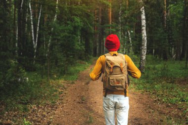 Sırt çantası ve kırmızı şapkalı bir turist karanlık ormanda ağaçların arasında yürüyor. Aktif yürüyüş ve doğa ile birlik kavramı.
