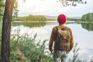 Sırt çantası ve kırmızı şapkalı bir turist gün batımında ormanın sularından güzel bir yansımayla nehrin kenarında duruyor. Aktif eğlenceyi ve doğayla bütünlüğü kavrayın