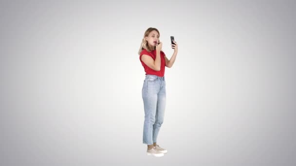 Mujer usando lápiz labial y mirando en su teléfono en el fondo de gradiente . — Vídeos de Stock