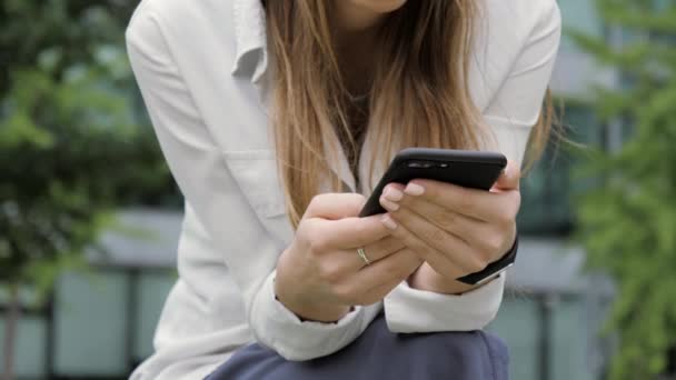 Vrouw in haar telefoon zittend op het bankje in het park te typen van een bericht. Close-up handen. — Stockvideo