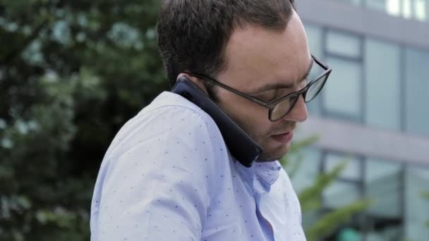 Hombre joven guapo vestido casualmente pasar tiempo al aire libre utilizando el ordenador portátil mientras está sentado en un banco y el uso de teléfono móvil . — Vídeo de stock