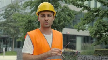 Hard Hat ve Safety Jacket Giyen Erkek Endüstriyel Mühendis Modern Binalarda Yürüyor ve Kamerayla Konuşuyor.