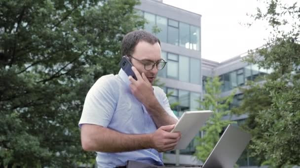 Uomo d'affari che utilizza tablet e laptop mentre parla su smartphone all'aperto . — Video Stock