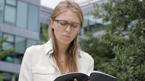 Joven estudiante con anteojos sentada en el banco al aire libre escribiendo notas en el cuaderno . — Vídeos de Stock