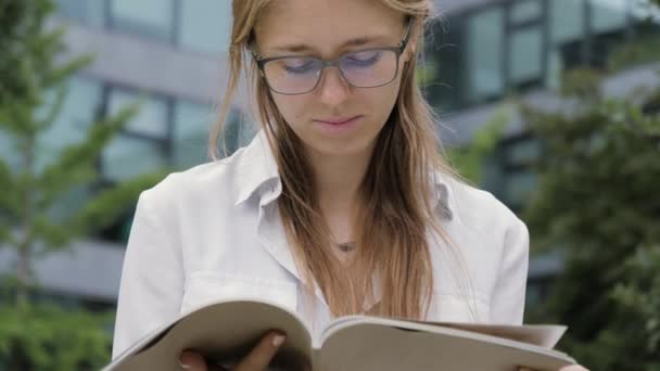 Een jonge blonde vrouw buiten in het park met een tijdschrift. — Stockvideo