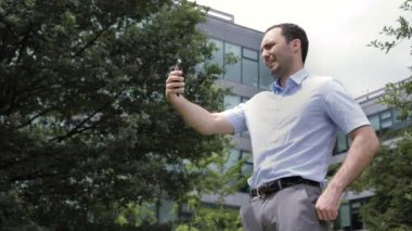 Neşeli yakışıklı genç adam gülümsüyor ve akıllı telefon kamerasıyla selfie çekiyor. Arka plandaki ofis binası..