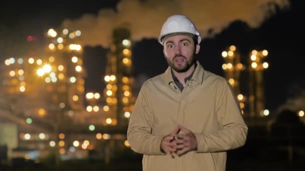 Молодий інженер носить білий твердий капелюх, пояснюючи щось на камеру, що стоїть біля заводу вночі . — стокове відео