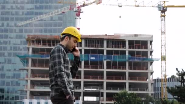 Ingenieur in een beschermende helm loopt en praat aan de telefoon op een bouwplaats. — Stockvideo