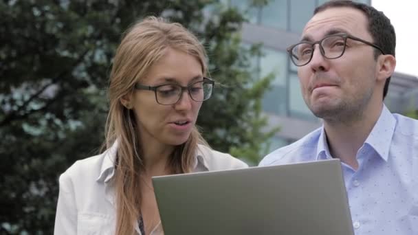 Aantrekkelijke man en vrouw zitten in het centrum van de stad in slimme casual business stijl samen te werken aan het project op laptop. — Stockvideo