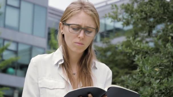 Young student girl wearing eyeglasses sitting on bench outdoors writing notes in notebook. — ストック動画