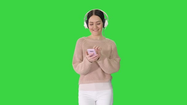 Glad flicka promenader lyssna på musik med smart telefon bär hörlurar på en grön skärm, Chroma Key. — Stockvideo