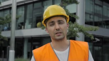 İnşaat işçisi modern binanın önünde kameraya konuşuyor..