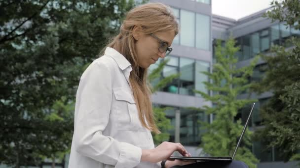 Jeune femme d'affaires utilisant un ordinateur portable dans un parc vert près de l'immeuble de bureaux . — Video