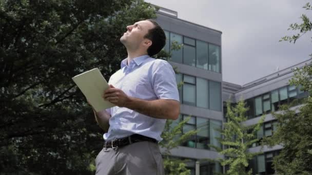 Бізнесмен стоїть в міському парку, біля офісних будівель, використовуючи планшет . — стокове відео