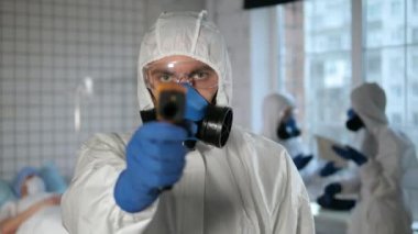 Koruyucu kıyafetli bir erkek doktor kameraya ısıyı ölçen dijital kızılötesi bir termometre tutuyor..