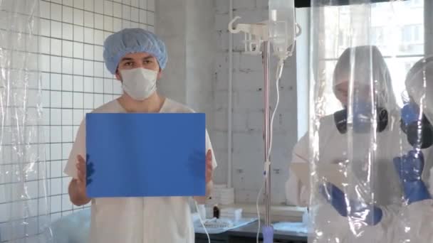 Patiënt houdt blanco bord en artsen in de beschermende pakken werken. — Stockvideo