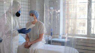 Koruyucu kıyafetli doktor hastanede tablet gösteren bir hastayla konuşuyor..