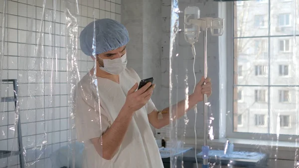 Paciente joven con una mascarilla médica usando teléfono en un hospital en cuarentena . —  Fotos de Stock