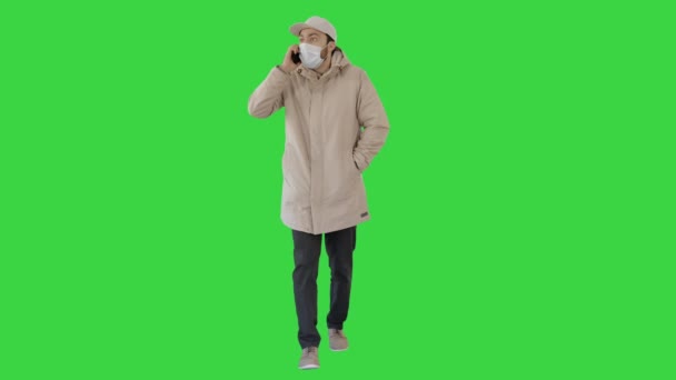 Casual man in outdoor kleding en medisch masker lopen en praten aan de telefoon op een groen scherm, Chroma Key. — Stockvideo