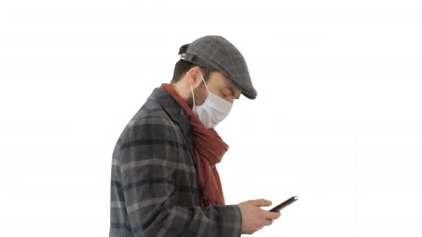 Стильний чоловік у медичній масці, що ходить та використовує смартфон на білому тлі . — стокове відео