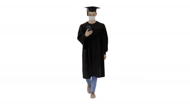 Joven con vestido de graduación caminando en máscara médica usando teléfono inteligente sobre fondo blanco . — Vídeo de stock