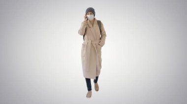 Koruyucu maskeli genç bir kadın gradyan arka planda cep telefonuyla yürüyor ve konuşuyor.