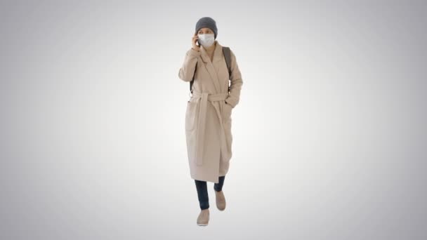 Молода жінка в захисній масці ходить і розмовляє мобільним телефоном на градієнтному фоні . — стокове відео