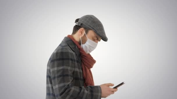 Стильный человек в медицинской маске ходьба и использование смартфона на градиентном фоне . — стоковое видео