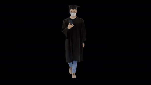 Jeune homme avec une robe de remise des diplômes marchant dans un masque médical à l'aide d'un smartphone, Alpha Channel — Video