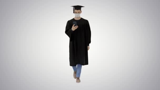 Young man with graduation gown walking in medical mask using smartphone on gradient background. — Stock Video
