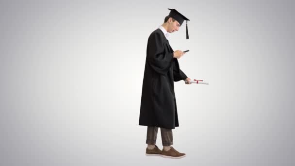 Graduación: Estudiante feliz caminando y mensajes de texto en su teléfono en el fondo gradiente . — Vídeos de Stock