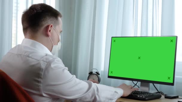 Biznesmen w masce medycznej spogląda poważnie na kamerę siedzącą obok pustego wyświetlacza. Wyświetlacz makiety z zielonym ekranem. — Wideo stockowe