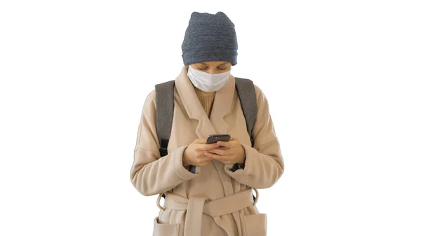Woman dressed in a coat wearing medical mask using phone and walking on white background. — Stock Photo, Image