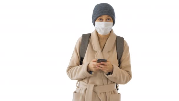 Woman dressed in a coat wearing medical mask using phone and walking on white background. — Stock Video