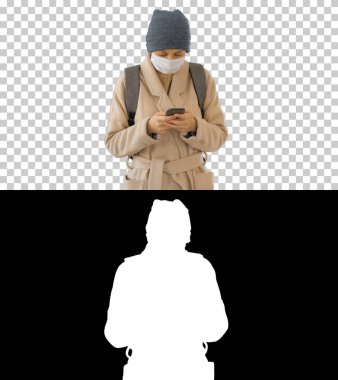 Paltolu bir kadın tıbbi maske takıyor telefon kullanıyor ve yürüyor, Alpha Channel 'da.