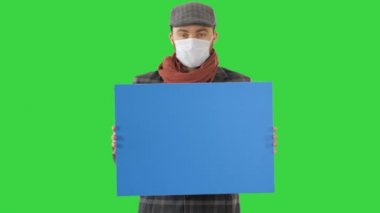 Moda kıyafetli ve tıbbi maskeli bir adam yeşil ekranda boş bir levha tutuyor, Chroma Key..