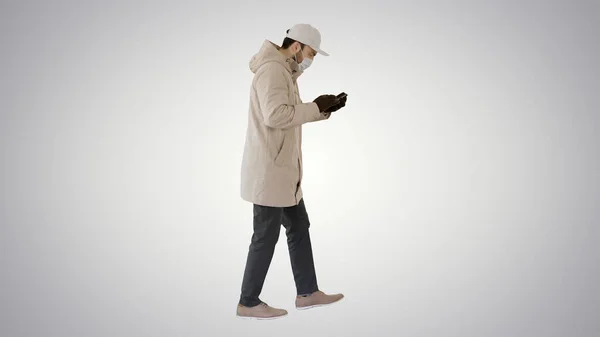 Homme caucasien dans un masque médical marche et en utilisant le téléphone sur fond de gradient . — Photo