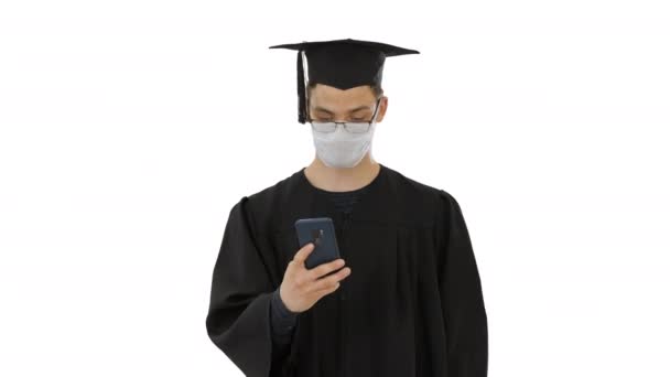 Junge raduate Student in medizinischer Maske mit Telefon zu Fuß auf weißem Hintergrund. — Stockvideo
