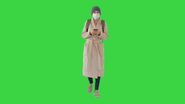 Γυναίκα ντυμένη με παλτό φορώντας ιατρική μάσκα χρησιμοποιώντας τηλέφωνο και περπατώντας σε μια πράσινη οθόνη, Chroma Key. — Αρχείο Βίντεο