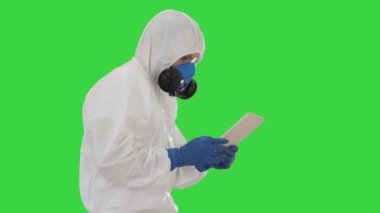 Bilimadamı ya da doktor biyo-tehlike giysileri ve koruyucu maskeler giyip yeşil ekranda yürürken dijital tablet kullanıyor..