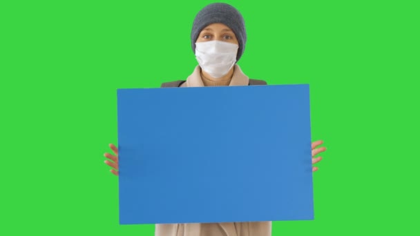 Donna in abiti da esterno e in maschera medica tenendo bordo vuoto su uno schermo verde, Chroma Key . — Video Stock