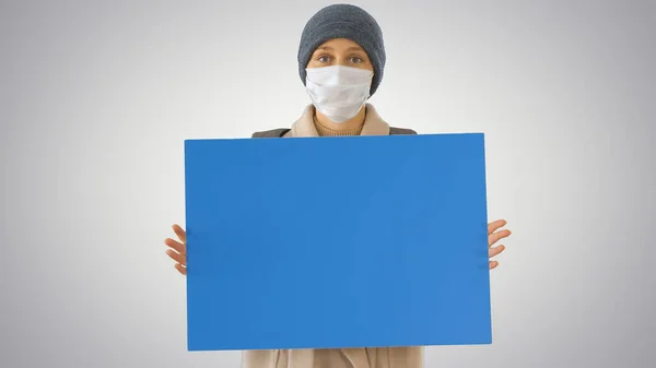 Mujer en ropa de exterior y en máscara médica sosteniendo tablero en blanco sobre fondo degradado . — Foto de Stock