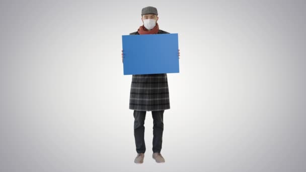 Man i en trendig outfit och medicinsk mask håller tomma plakat på lutning bakgrund. — Stockvideo