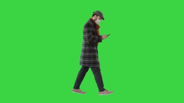 Mann mit medizinischer Maske geht und benutzt Mobiltelefon auf einem Green Screen, Chroma Key. — Stockvideo