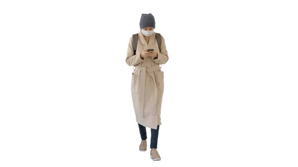 Vrouw gekleed in een jas met medisch masker met behulp van telefoon en lopen op een witte achtergrond. — Stockfoto